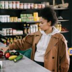 A woman selecting fresh fruits and vegetables at a grocery store with shelves stocked in the background.